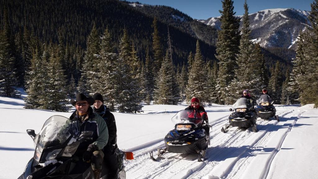 Four people ride snowmobiles through trees near Taos Ski Valley.